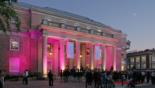 A an exterior view of a performing arts venue, lit up with pink lights in between its large columns.