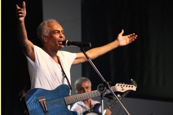 A woman wearing a white t-shirt sings with her arms outstretched.