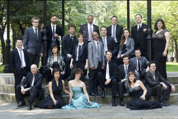 A group of people in suits and formal dresses sit for a formal group picture in a park.