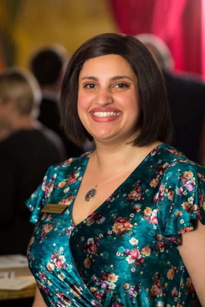 A woman wearing a green floral dress and Carolina Performing Arts name tag smiles while hosting a donor reception.