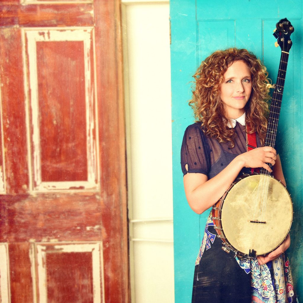 white woman stands with banjo in front of teal, white, and brown doors