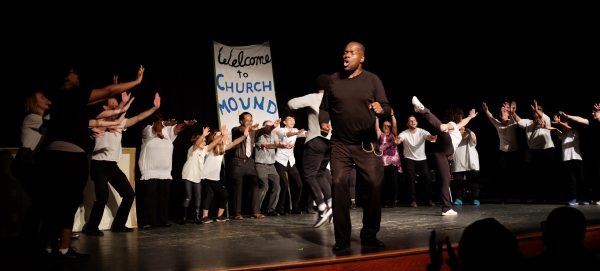 Black man dressed in black on foreground of stage, with several people in background holding up a hand-painted sign that says "Welcome to Church Mound"