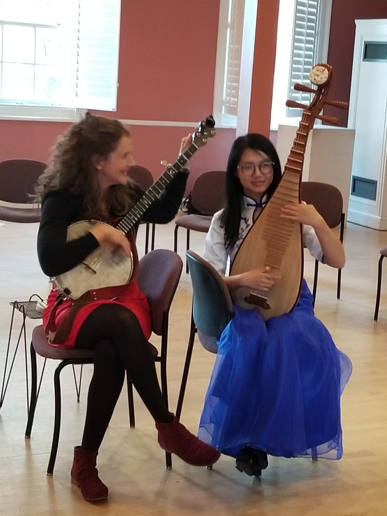 A white woman in black top and tights and red skirt plays the banjo seated next to a young Asian woman with a white top and long blue skirt, playing a Chinese lute.