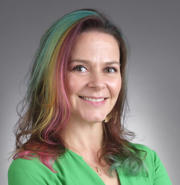 White woman with rainbow stripes on light brown hair, smiling and wearing green v-neck top and arms crossed