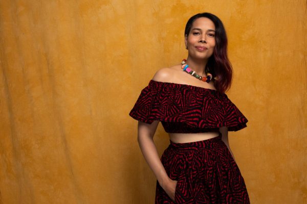Artist Rhiannon Giddens leans up against a tan wall, wearing a two-piece dress and a beaded necklace.