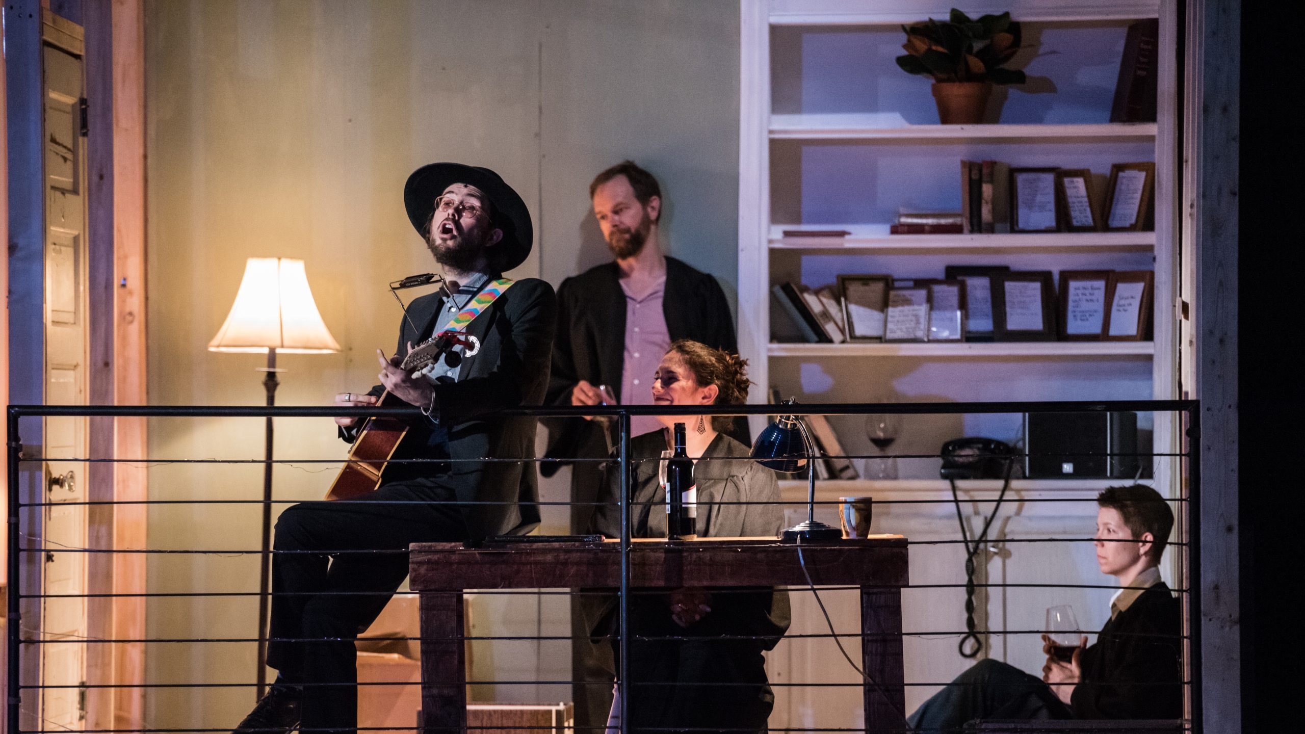 A man in a hat holds a guitar and sings, as three other people look on.