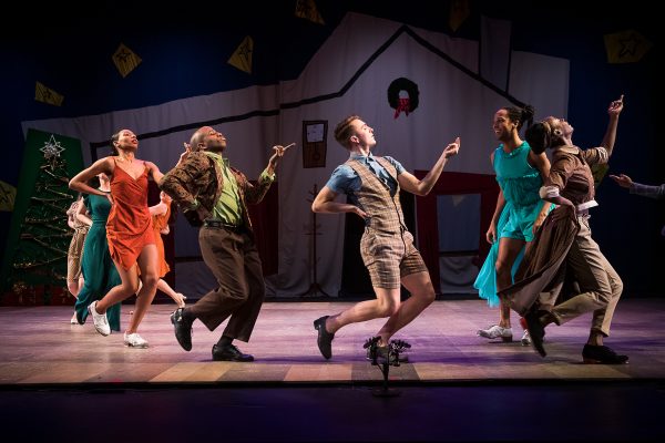 Dancers, attired in colorful dresses and suits, dance across the stage, fingers upraised.