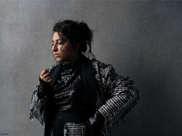 A woman stands against a gray background, dressed in gray and black.