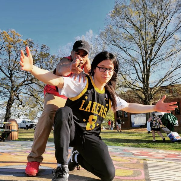 A man poses in a black hat, a red basketball jersey, a jacket, and red shoes. He is pointing at the camera, with his left hand on the shoulder of another man in a black Lakers jersey, whose arms are spread. The man in the Lakers jersey is kneeling.