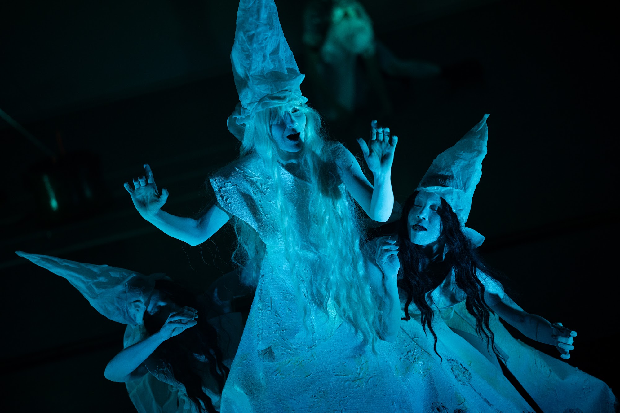 Demigods in white paint and tall sculptural headpieces reach outward in blue-green light
