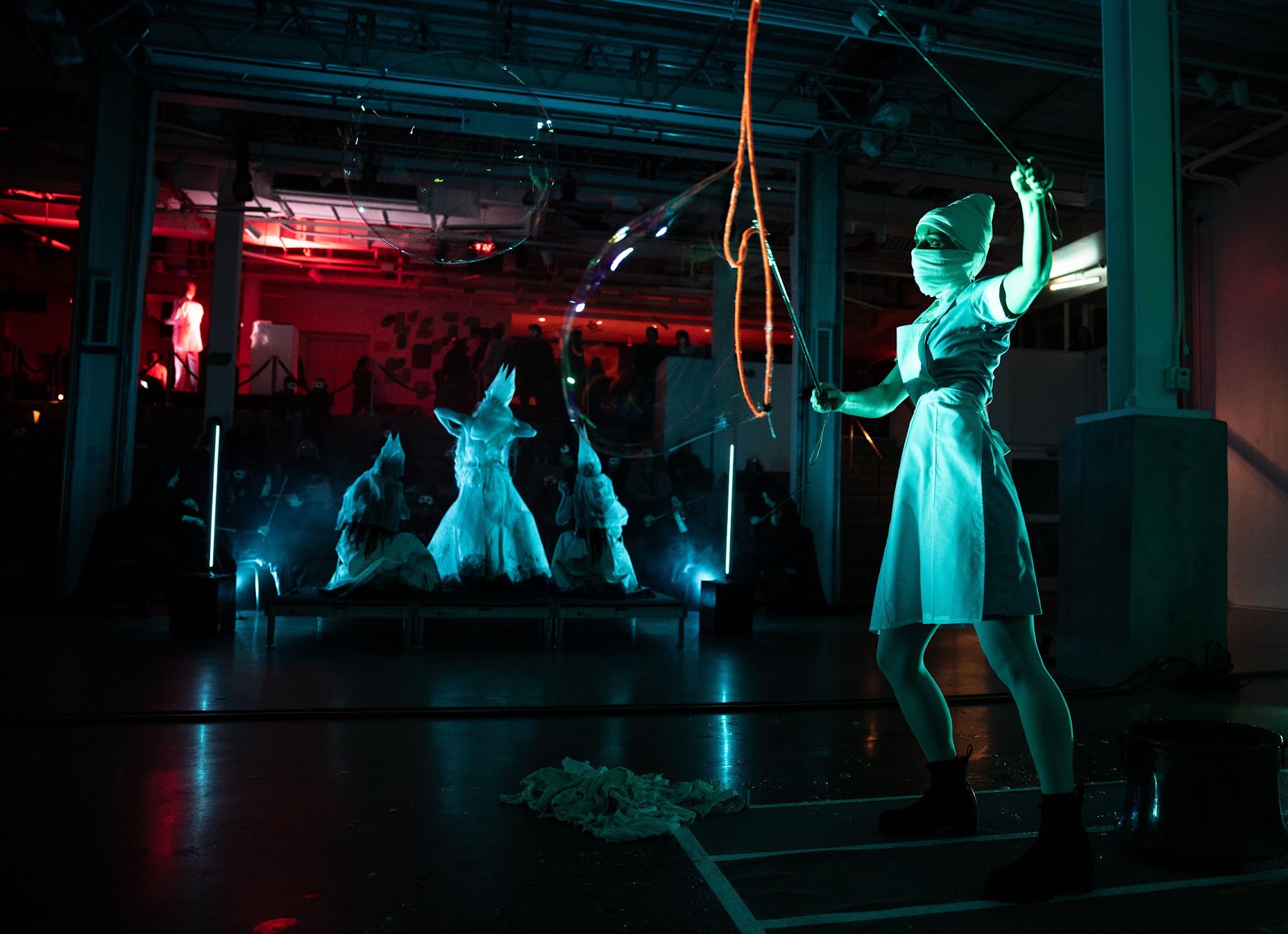 A nurse in a vintage uniform reaches toward a hanging rope, with white-costumed demigods posed behind her in teal light