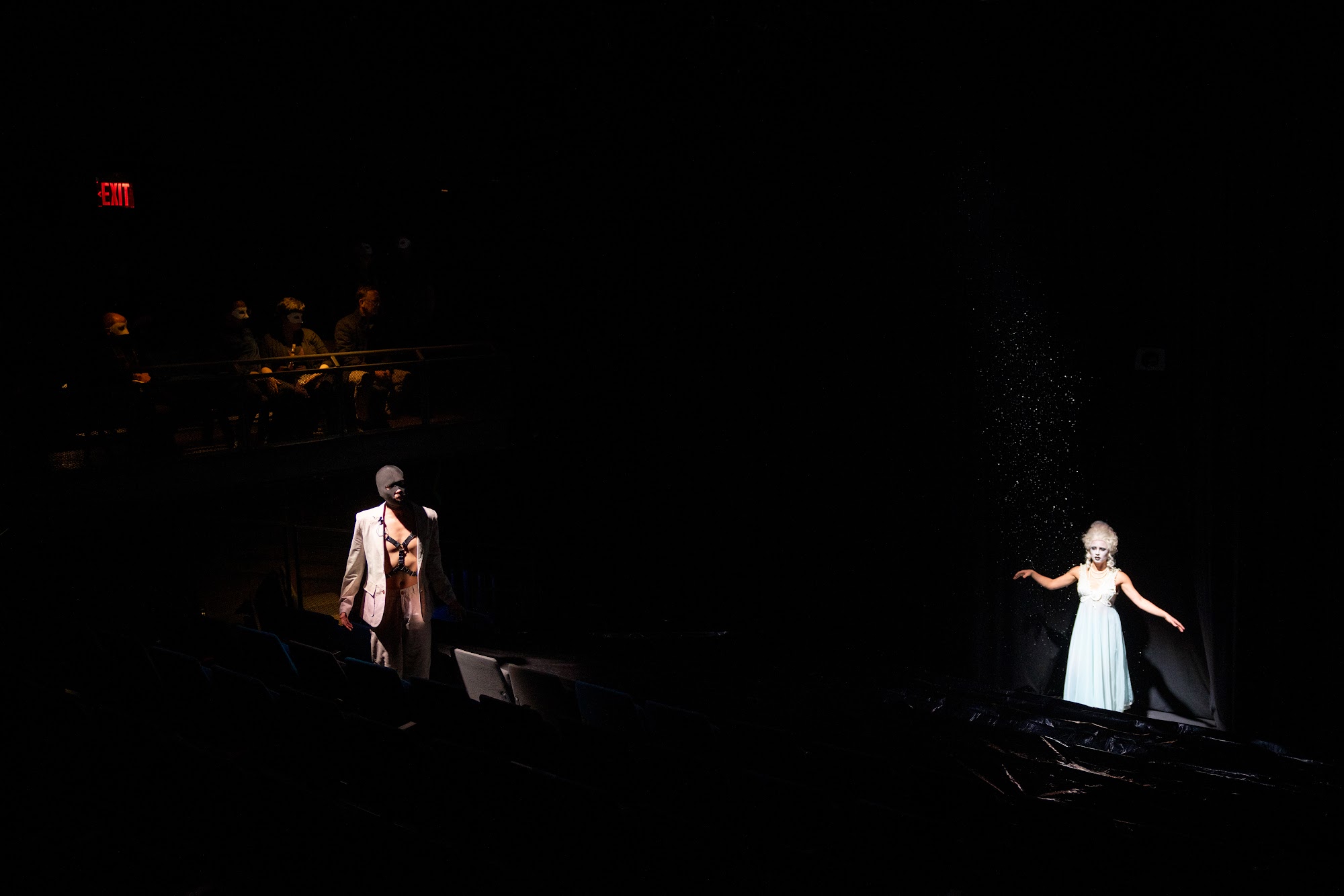 The Snow Dancer in a pale dress stands in a pool of light in a dark room as another figure watches